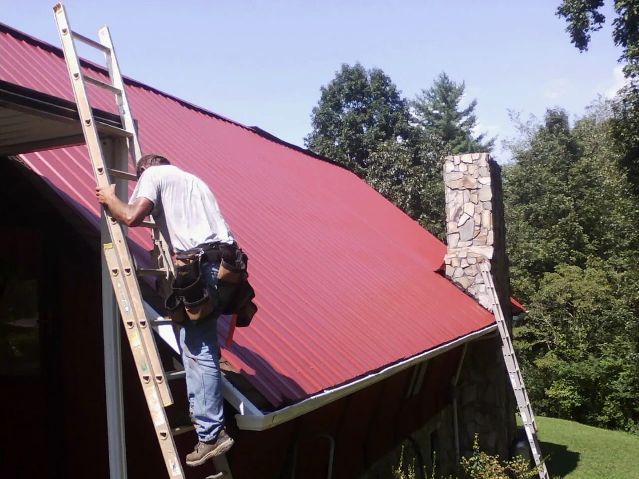 Roofer on a ladder beside a red metal roof and stone chimney — Soffit & Fascia Repair by Roofing Brothers in South Acworth